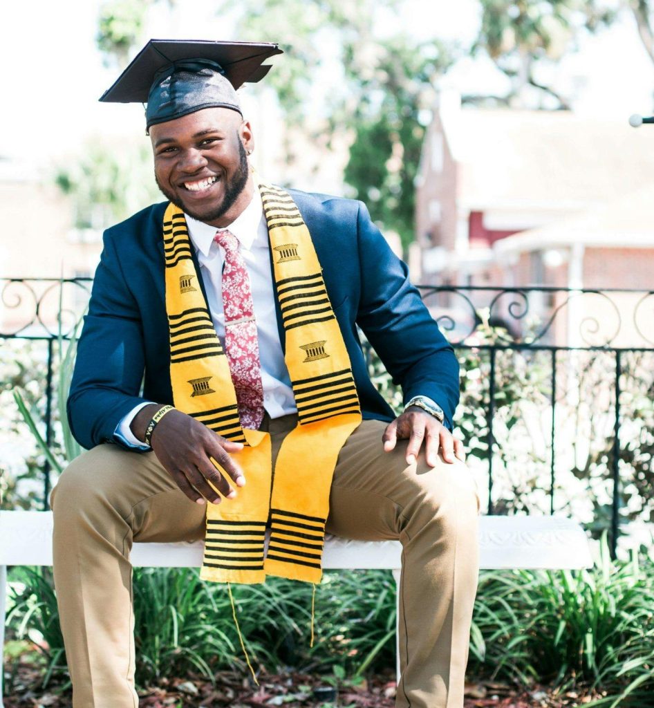 Smiling graduate sitting outside wearing a cap and gown with stole.
